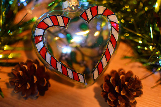 Heart-shaped candy cane ornament with red and white stripes on a wooden surface with pine cones and greenery.